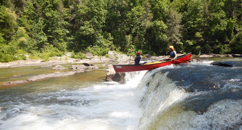 Two people wearing safety gear navigate a canoe over a short waterfall. Behind them is a rocky shore and dense trees.